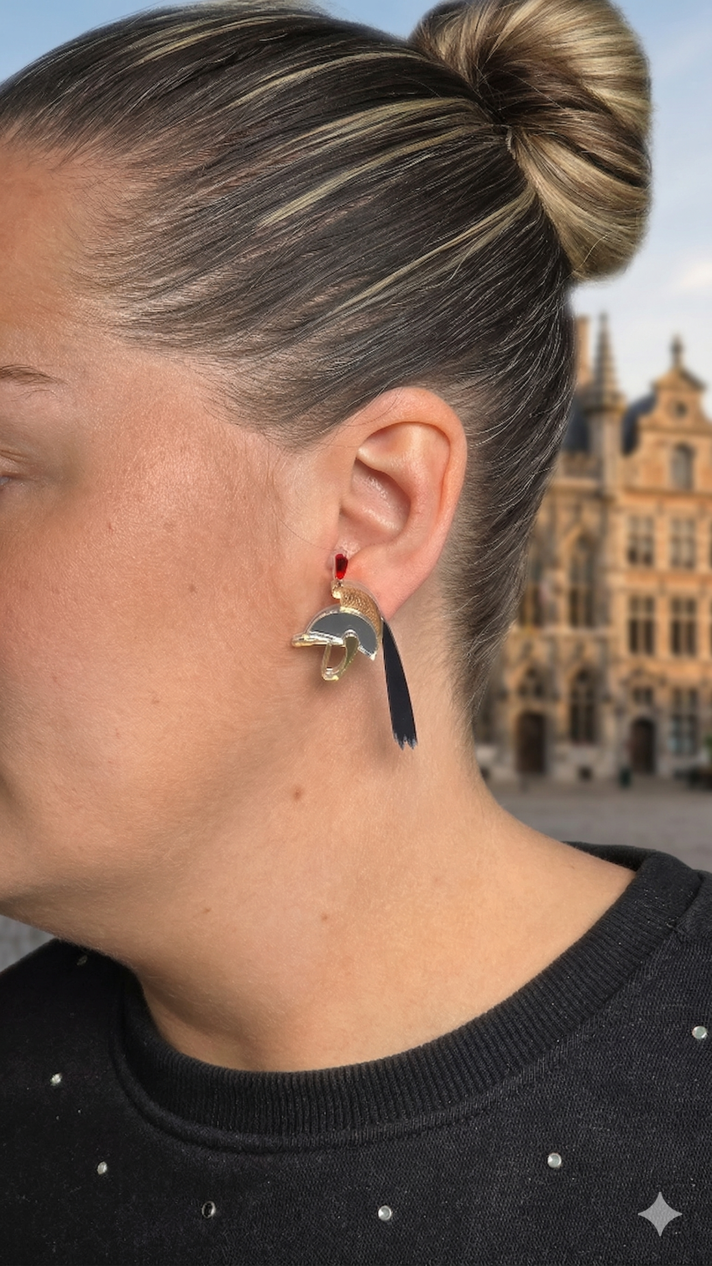 Portrait de profil d'une femme portant une boucle d'oreille unique de la marque Léawood. Le bijou est une création géométrique stylisée en forme d'oiseau, réalisée avec des segments d'acrylique miroir de couleur or, noir et gris fumé, reliés par un petit crochet rouge. L'arrière-plan flouté montre les bâtiments historiques de la Grand-Place de Mons, en Belgique. La boucle d'oreille est le point focal de l'image.